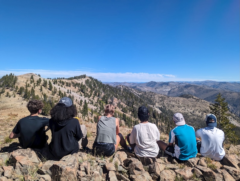 4 people sit on a rocky outcrop admiring the view from high up on a peak